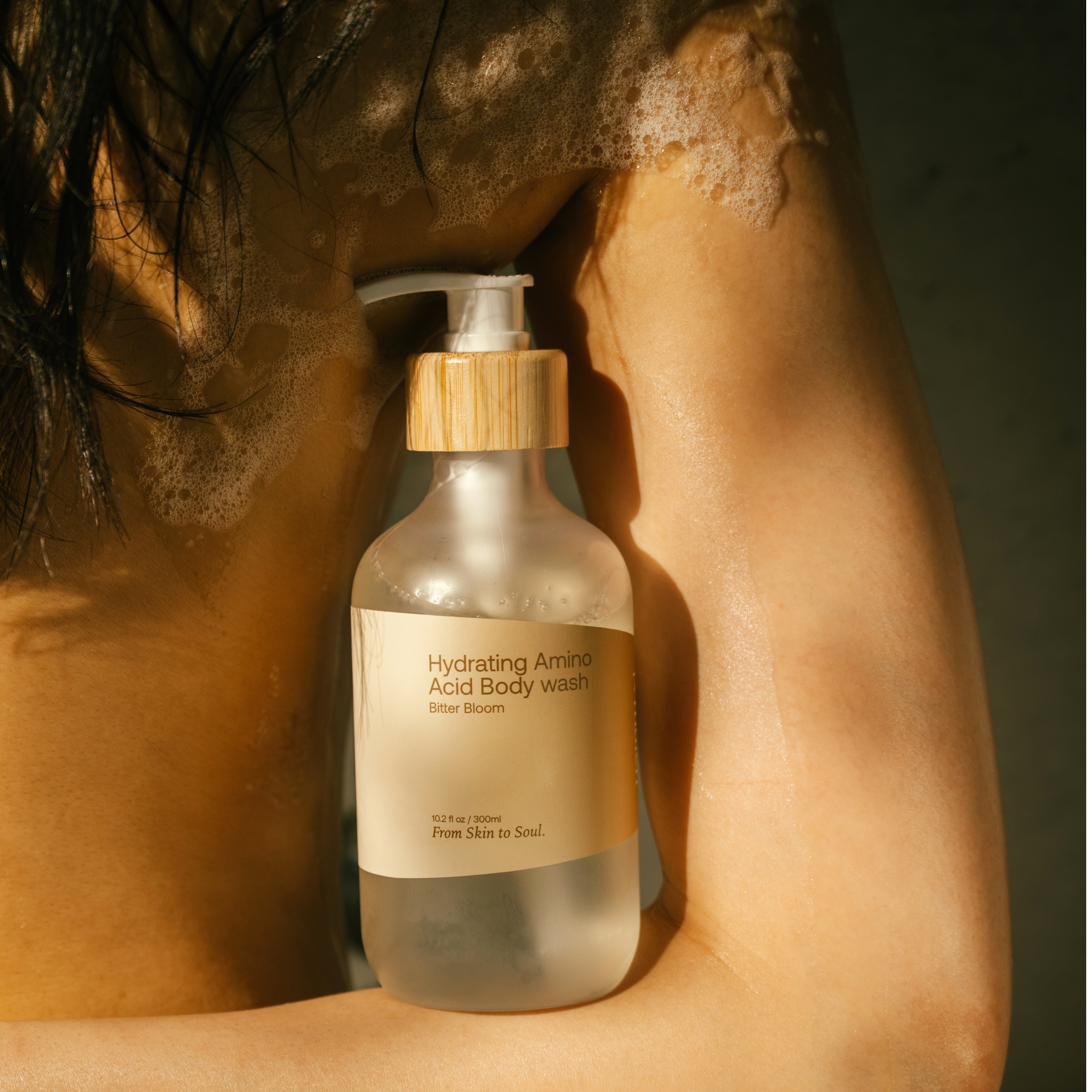 Bottle of 'Hydrating Amino Acid Body Wash' held against a person's skin with soap suds.